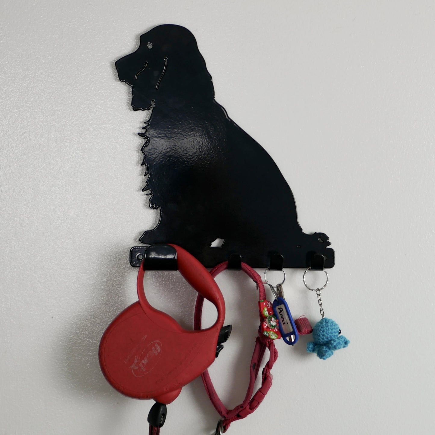 A close shot of a black cocker spaniel dog key rack with one large hook and three smaller hooks along the bottom. Hanging from the hooks are a red dog lead, red dog collar, a set of keys, and a keyring.