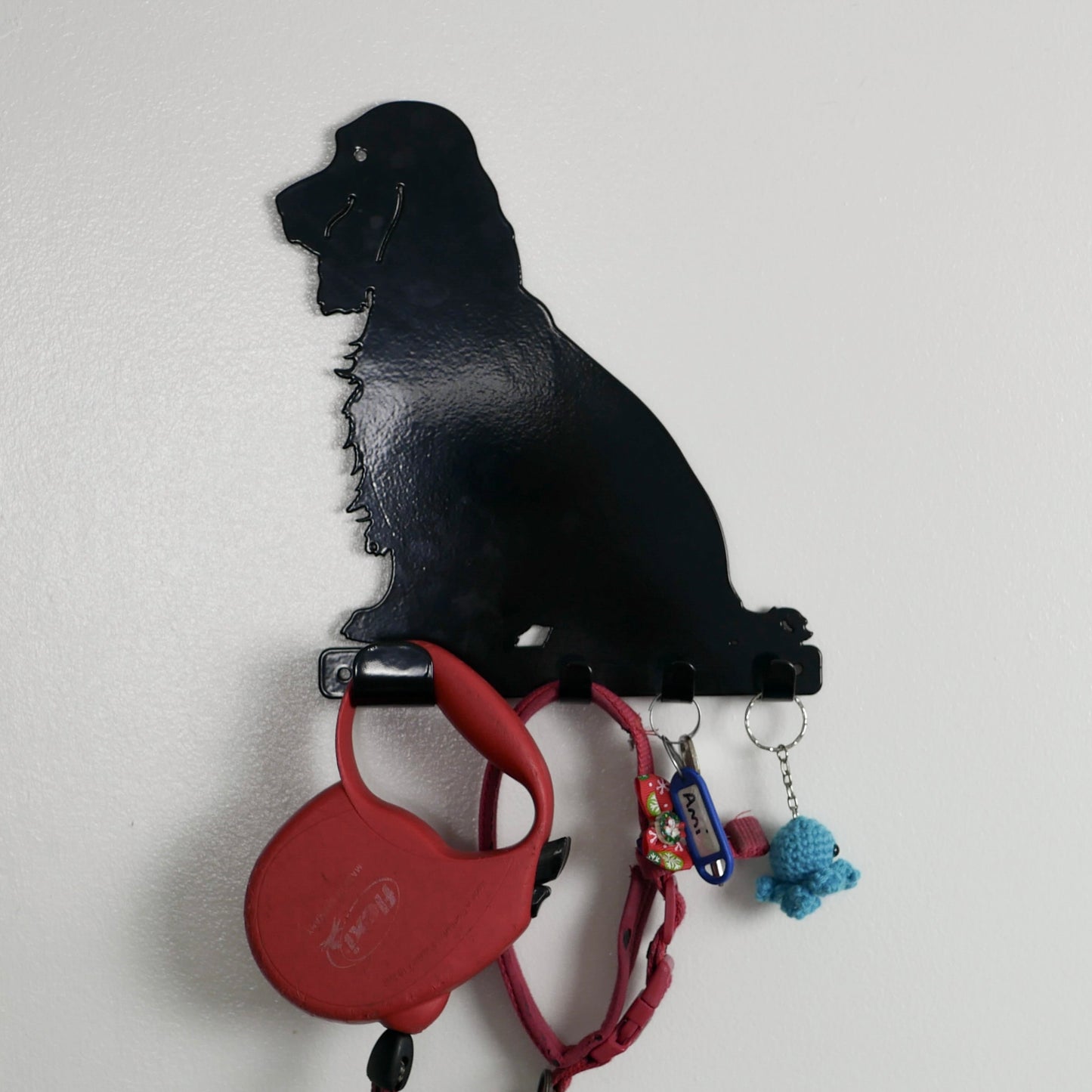 A close shot of a black cocker spaniel dog key rack with one large hook and three smaller hooks along the bottom. Hanging from the hooks are a red dog lead, red dog collar, a set of keys, and a keyring.