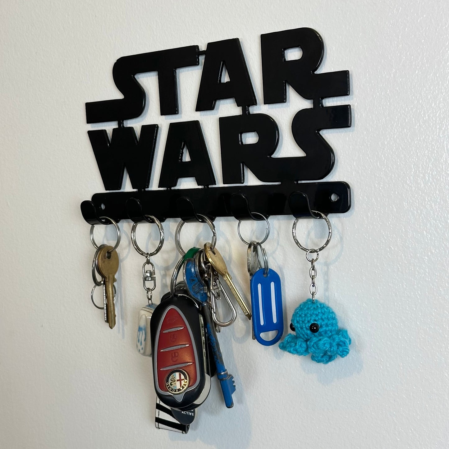 A close up photo of a black Star Wars logo hanging key rack with 5 small hooks. Hanging from the hooks are two sets of keys, a car key, and two keyrings.