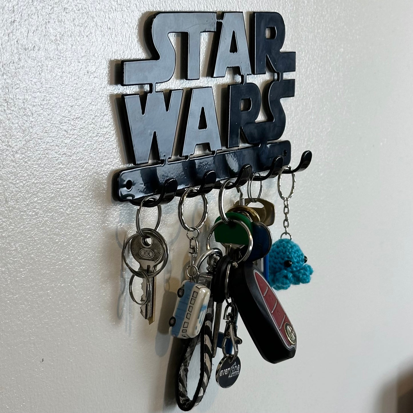 A left side view of a black Star Wars logo hanging key rack with 5 small hooks. Hanging from the hooks are two sets of keys, a car key, and two keyrings.