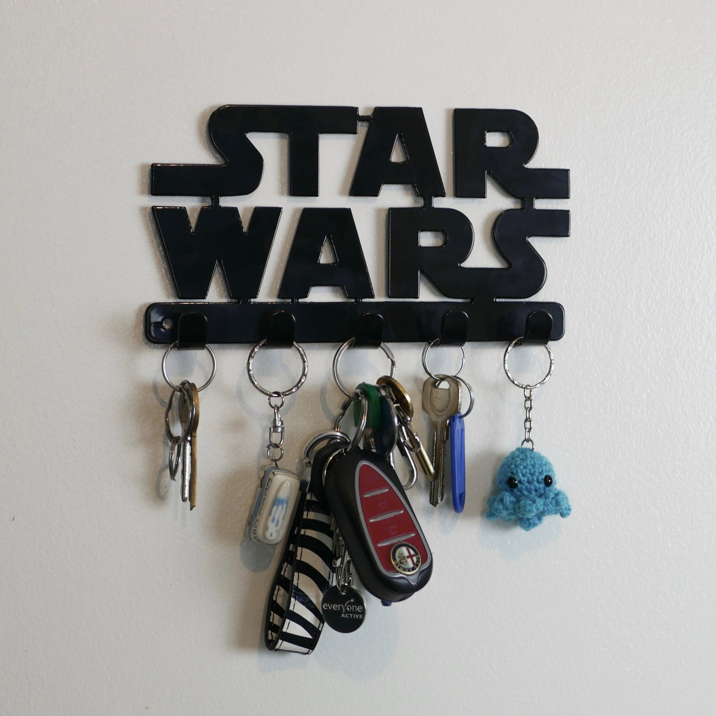 A black Star Wars logo hanging key rack with 5 small hooks. Hanging from the hooks are two sets of keys, a car key, and two keyrings.
