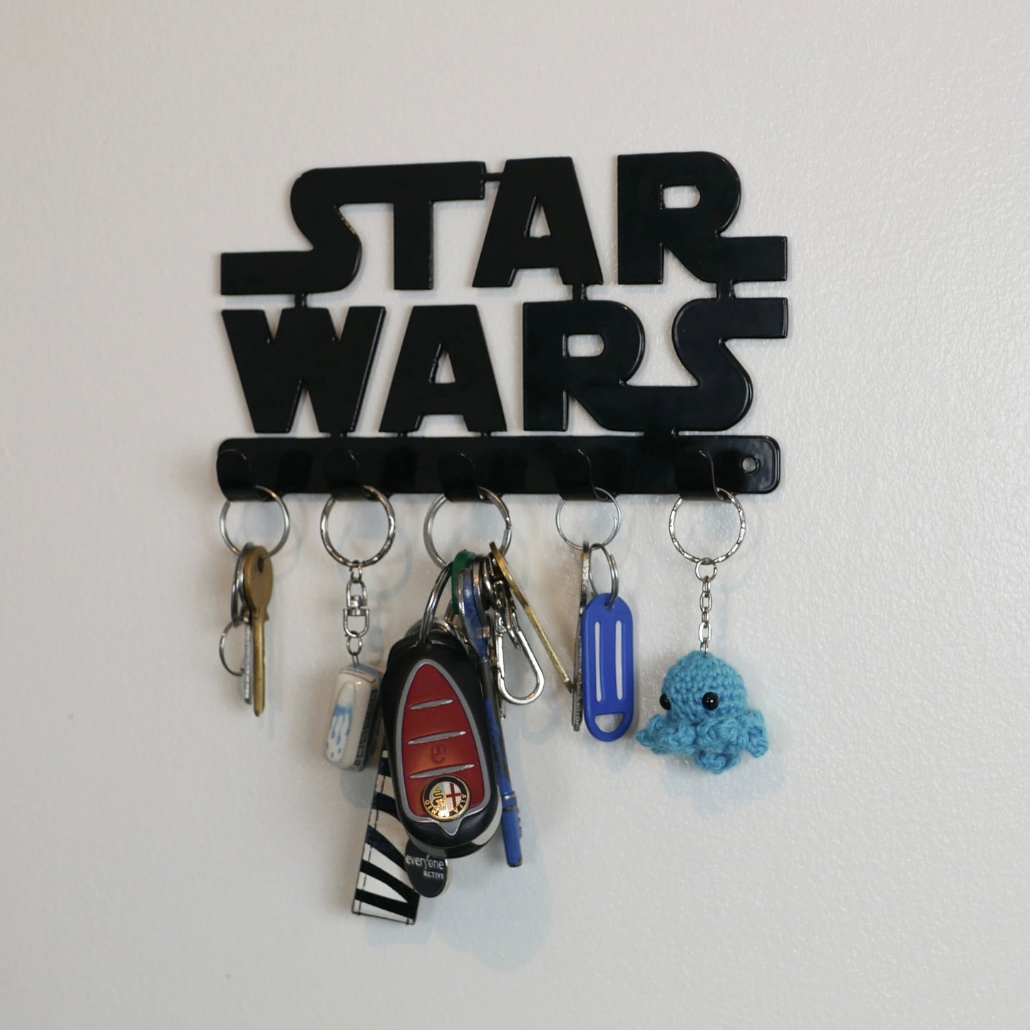 A right side view of a black Star Wars logo hanging key rack with 5 small hooks. Hanging from the hooks are two sets of keys, a car key, and two keyrings.