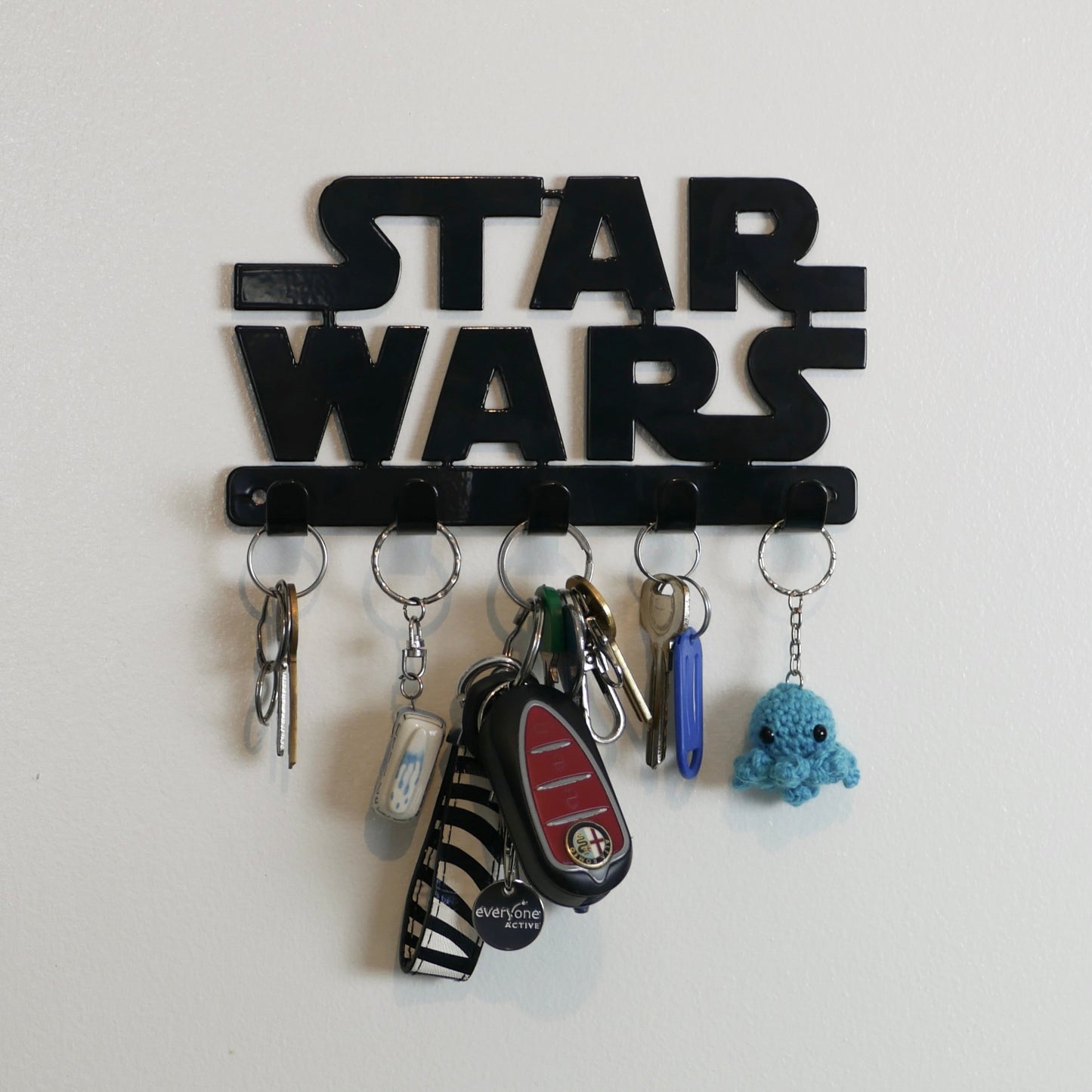 A black Star Wars logo hanging key rack with 5 small hooks. Hanging from the hooks are two sets of keys, a car key, and two keyrings.