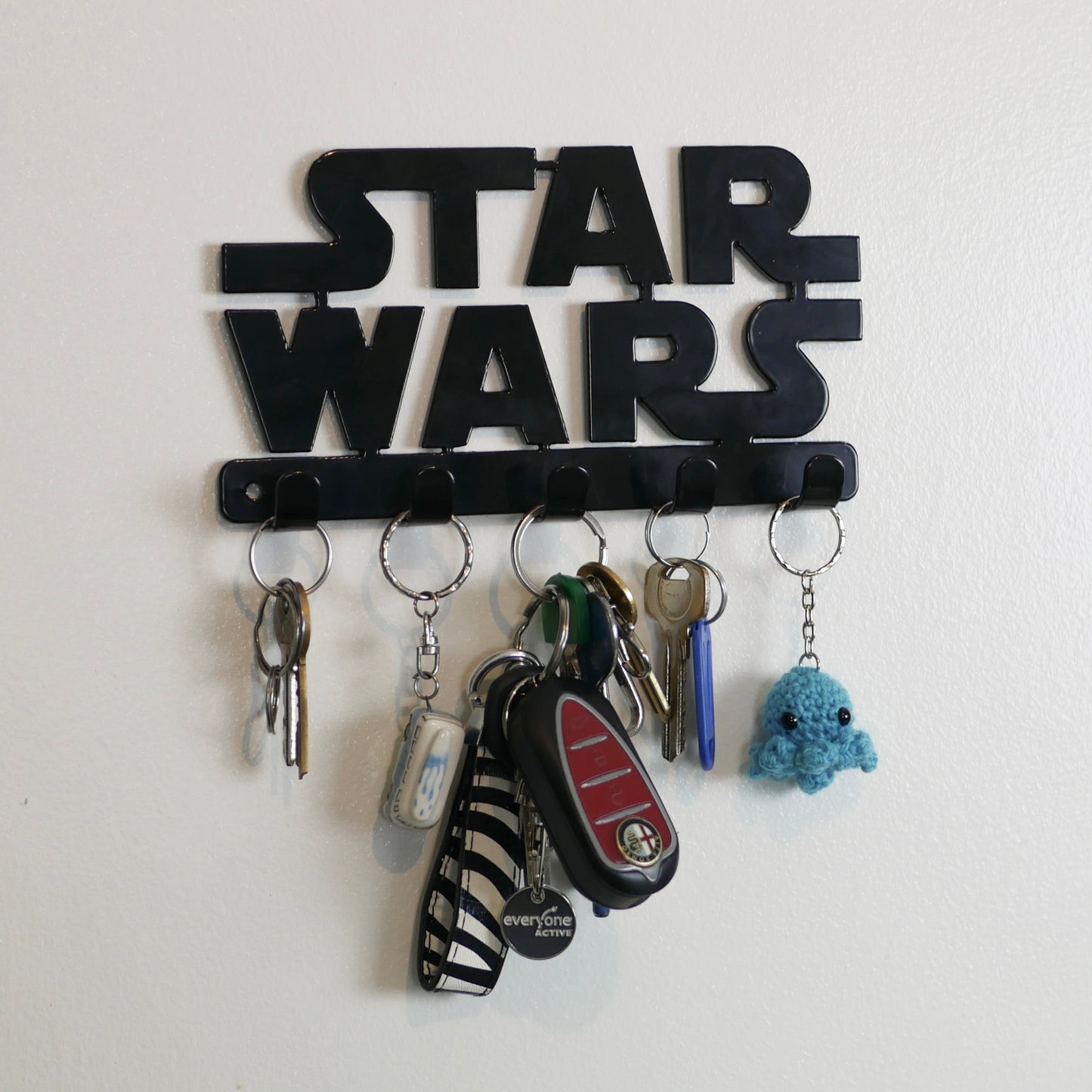 A black Star Wars logo hanging key rack with 5 small hooks. Hanging from the hooks are two sets of keys, a car key, and two keyrings.