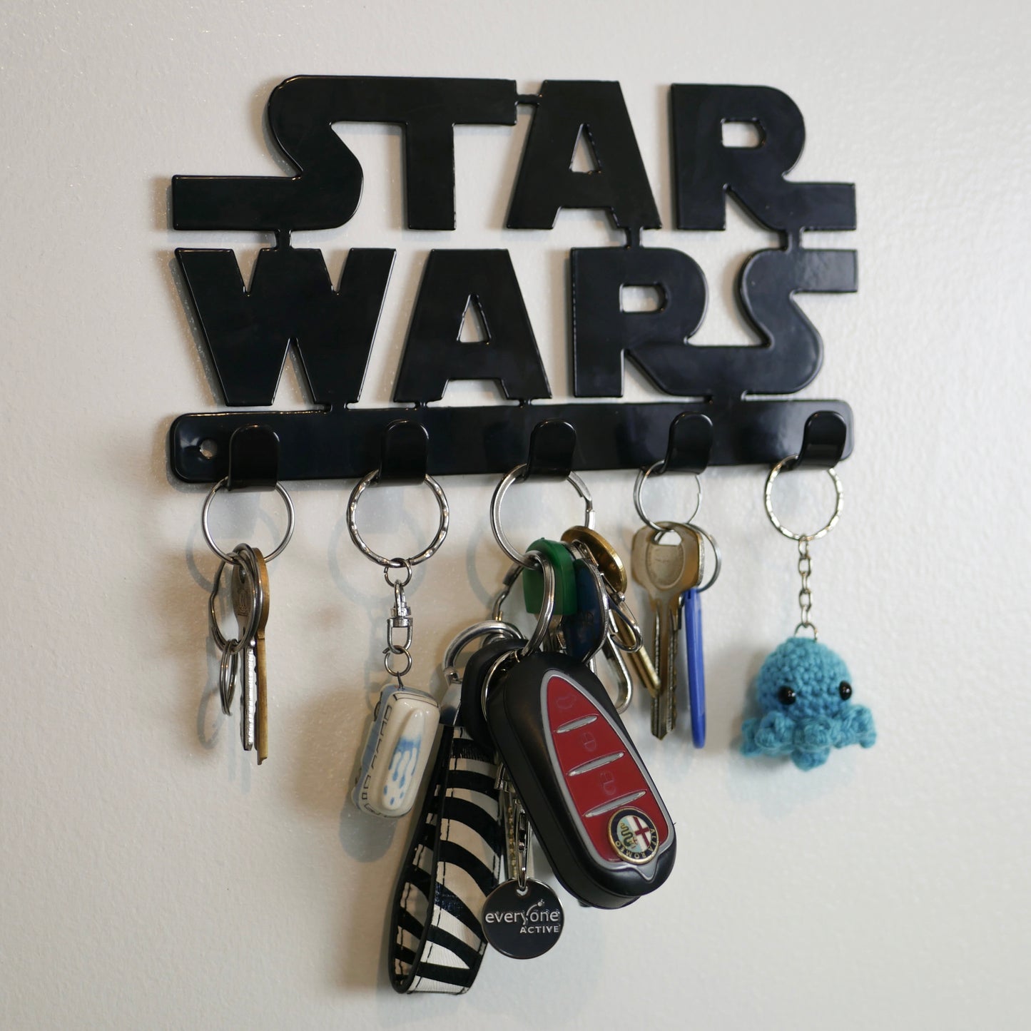A black Star Wars logo hanging key rack with 5 small hooks. Hanging from the hooks are two sets of keys, a car key, and two keyrings.