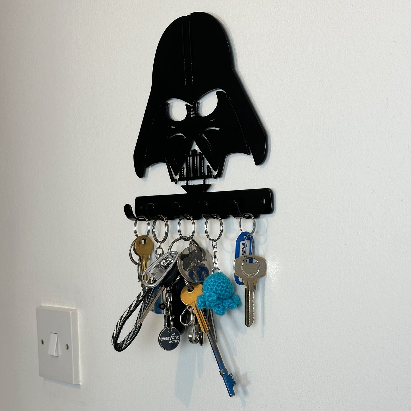 A right side view of a black metal Star Wars Darth Vader hanging plaque with five small hooks along the bottom. Hanging from these hooks are two sets of keys, a set of car keys, and two keyrings.