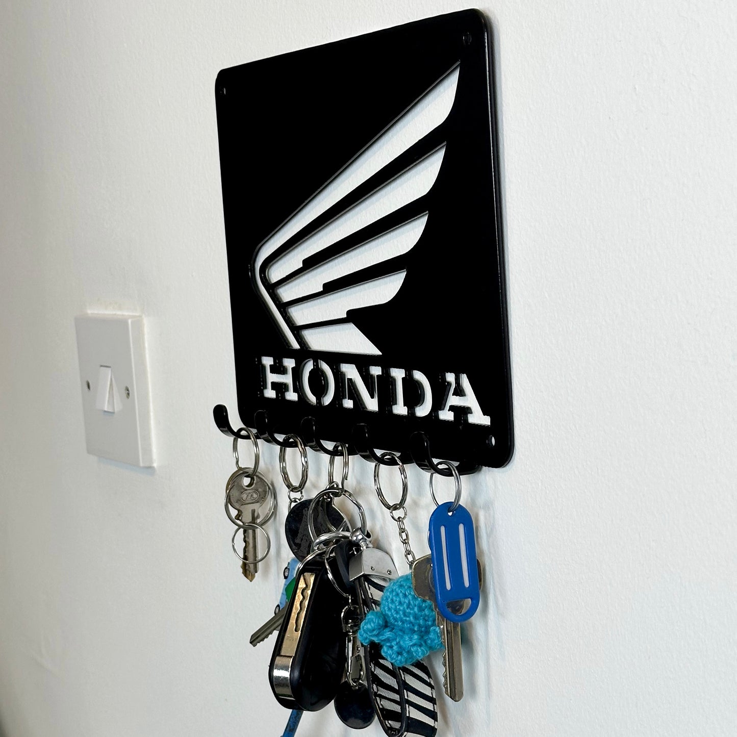 A side view of a black metal Honda logo hanging plaque with five small hooks along the bottom. Hanging from these hooks are two sets of keys, a set of car keys, and two keyrings.
