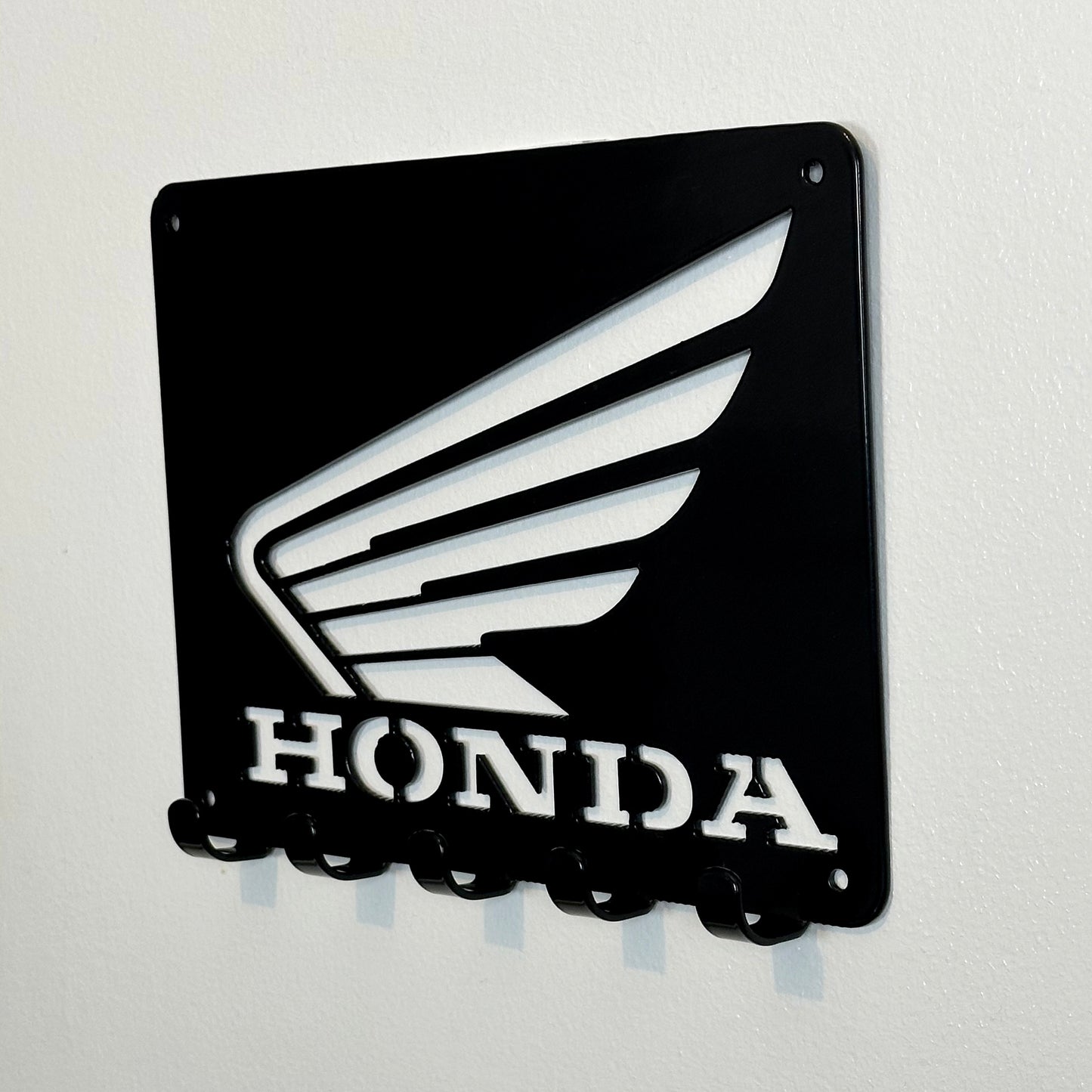 A side view of a black metal Honda logo hanging plaque with five small hooks along the bottom.