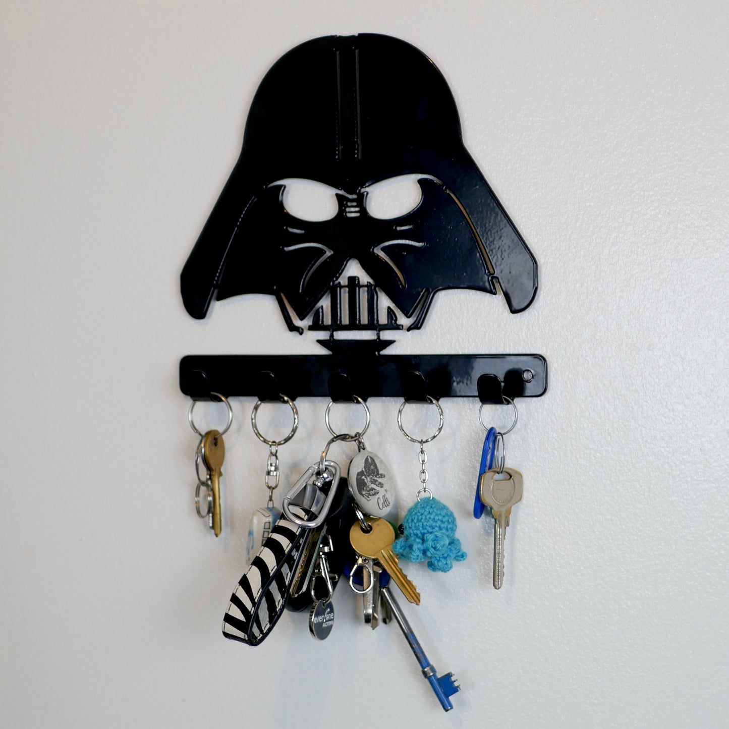 A black metal Star Wars Darth Vader hanging plaque with five small hooks along the bottom. Hanging from these hooks are two sets of keys, a set of car keys, and two keyrings.