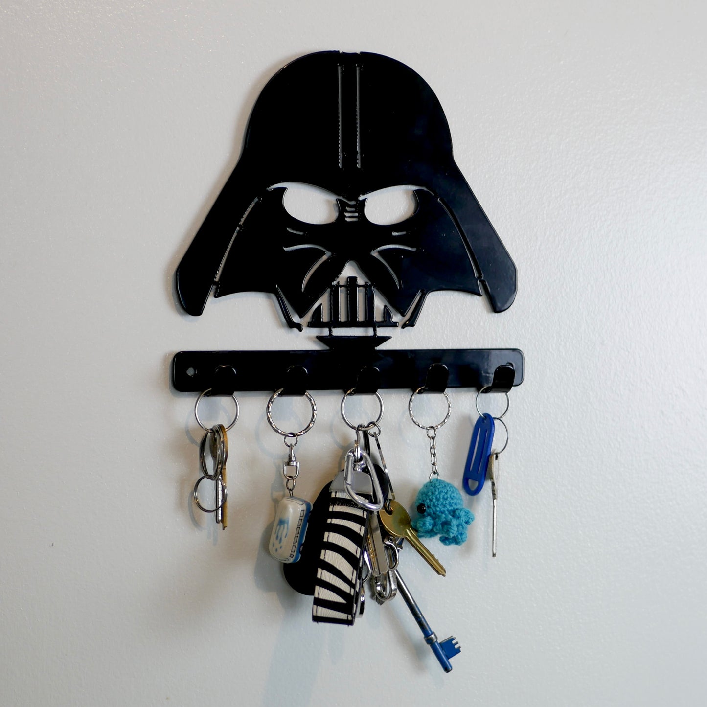 A black metal Star Wars Darth Vader hanging plaque with five small hooks along the bottom. Hanging from these hooks are two sets of keys, a set of car keys, and two keyrings.