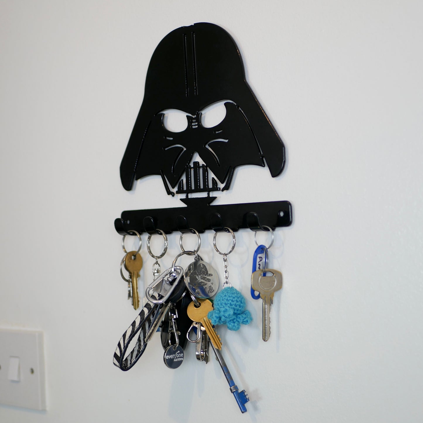 A right side view of a black metal Star Wars Darth Vader hanging plaque with five small hooks along the bottom. Hanging from these hooks are two sets of keys, a set of car keys, and two keyrings.
