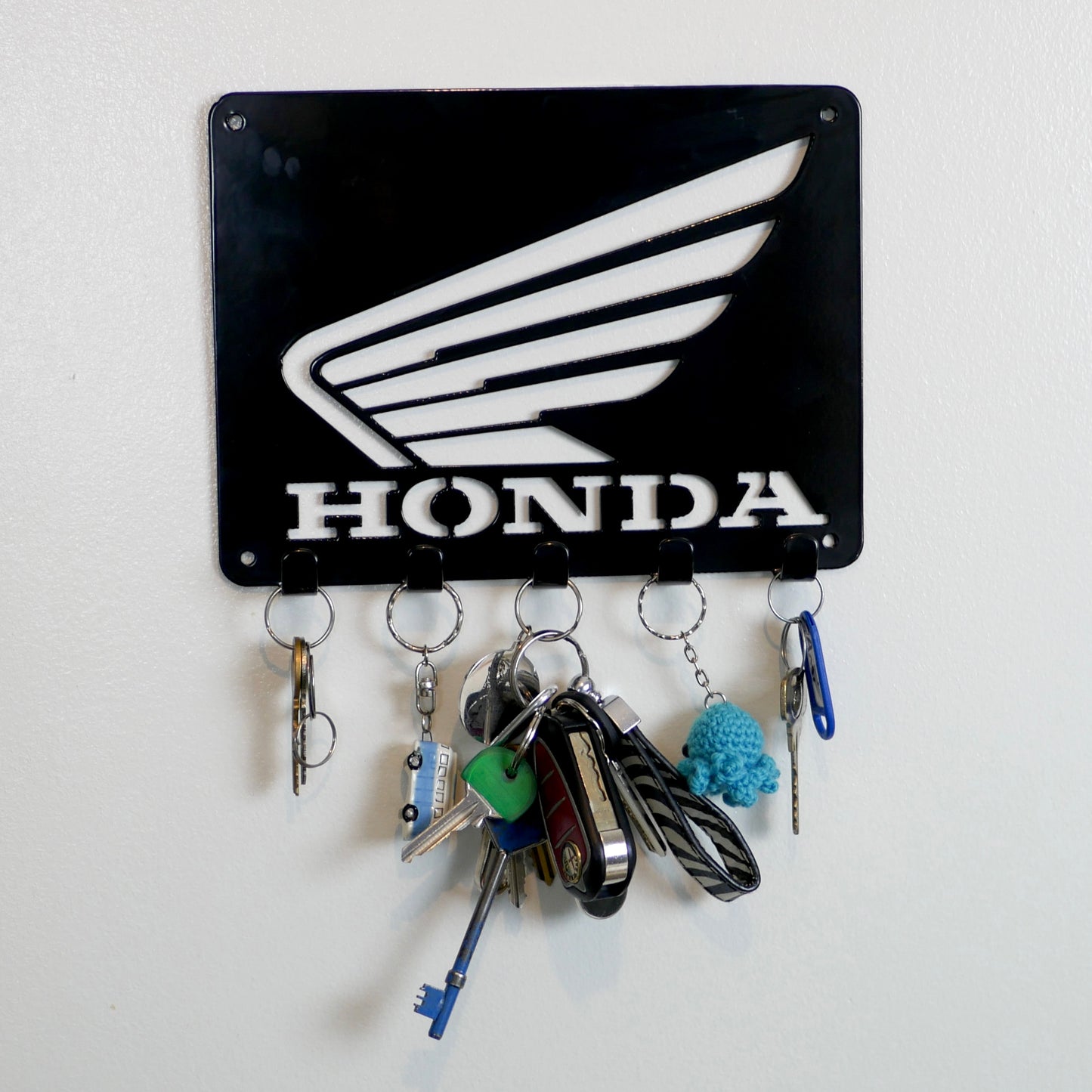 A black metal Honda logo hanging plaque with five small hooks along the bottom. Hanging from these hooks are two sets of keys, a set of car keys, and two keyrings.