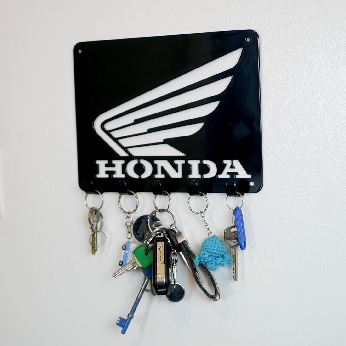 A black metal Honda logo hanging plaque with five small hooks along the bottom. Hanging from these hooks are two sets of keys, a set of car keys, and two keyrings.
