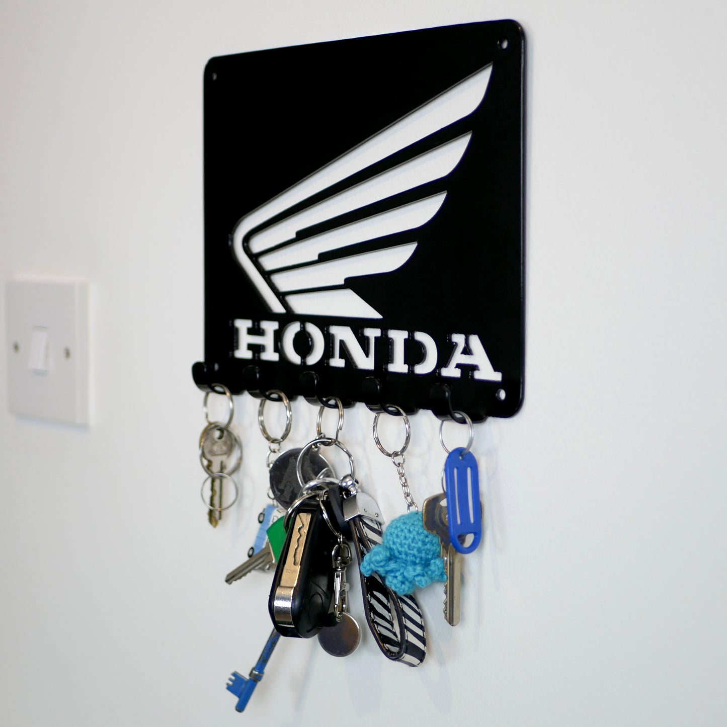 A right side view of black metal Honda logo hanging plaque with five small hooks along the bottom. Hanging from these hooks are two sets of keys, a set of car keys, and two keyrings.
