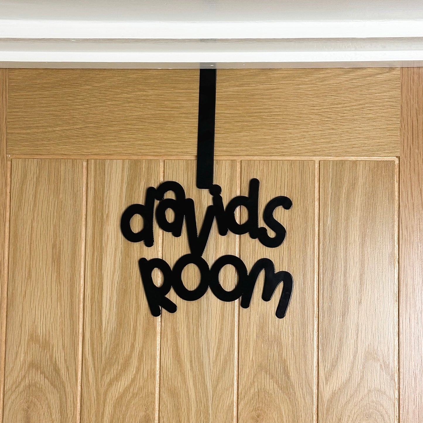 A wooden door with a black sign reading "davids room" that is in a cursive font. It is hung over the top of the door using an over door hook.