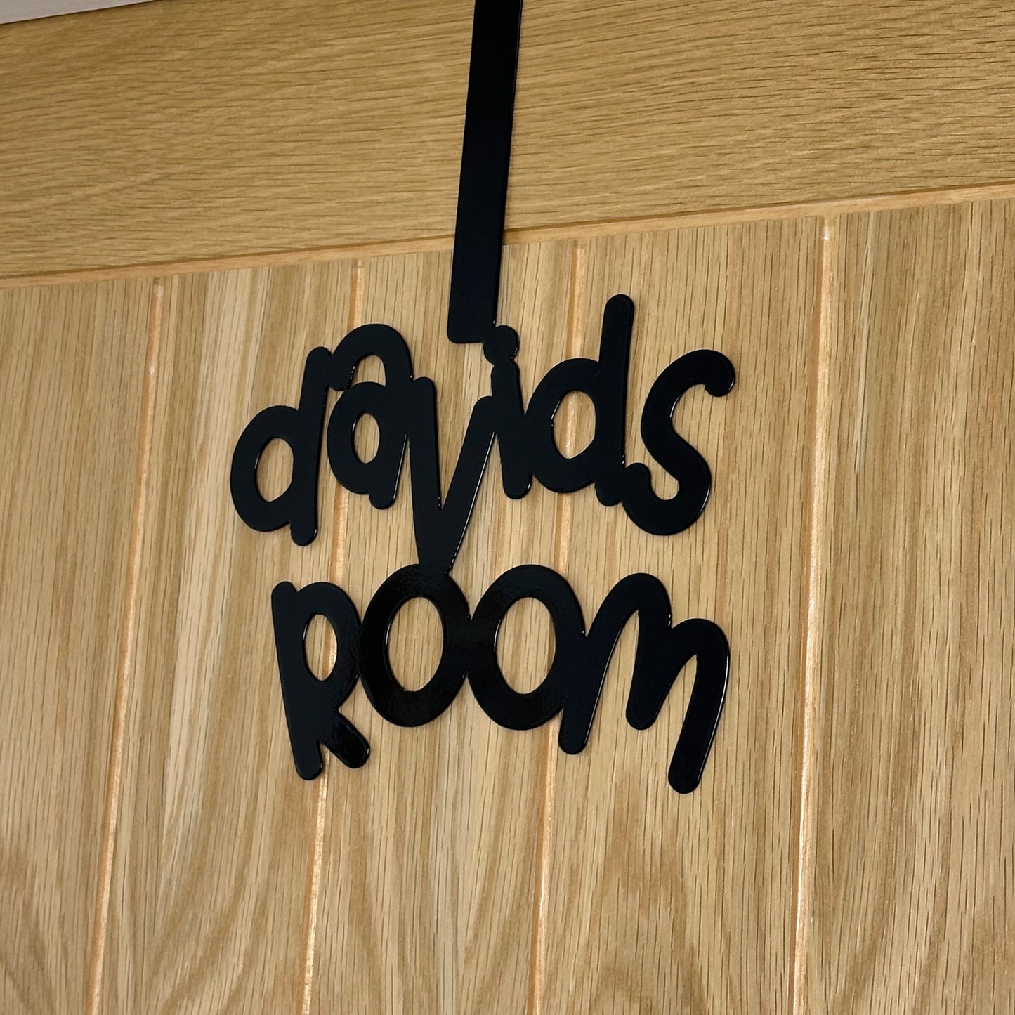 A side view of a wooden door with a black sign reading "davids room" in a cursive font. It is hung using an over door hook.