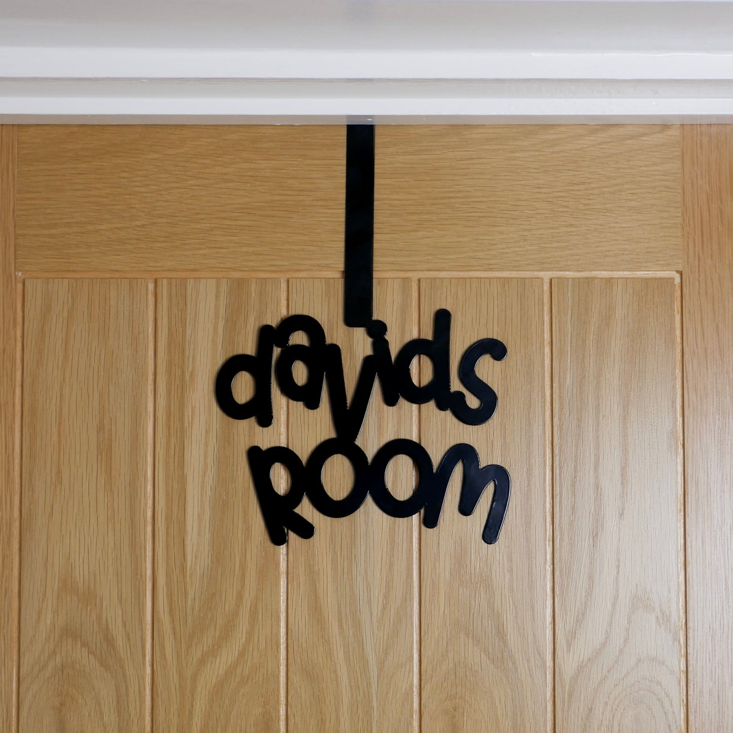A wooden door with a black sign reading "davids room" that is in a cursive font. It is hung over the top of the door using an over door hook.