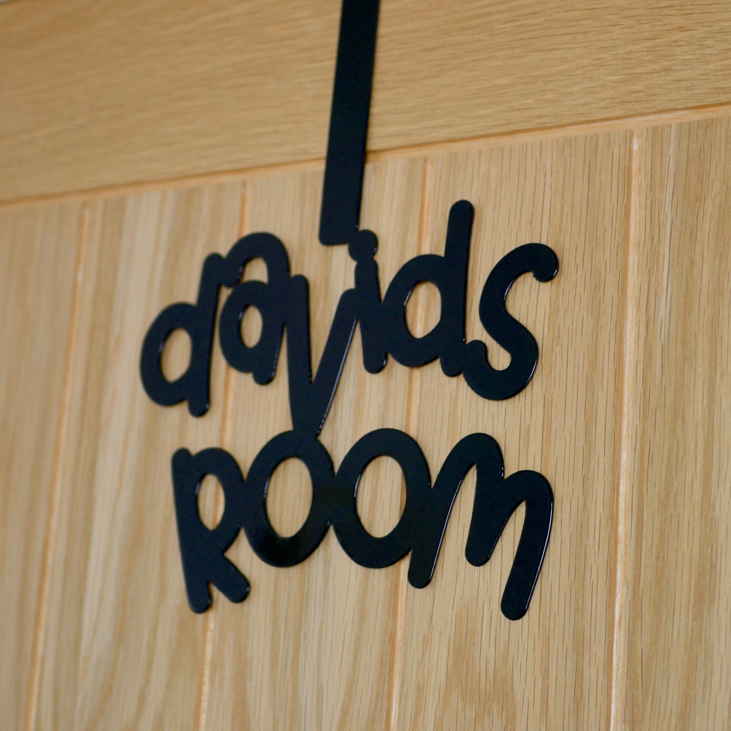 A side view of a wooden door with a black sign reading "davids room" in a cursive font. It is hung using an over door hook.
