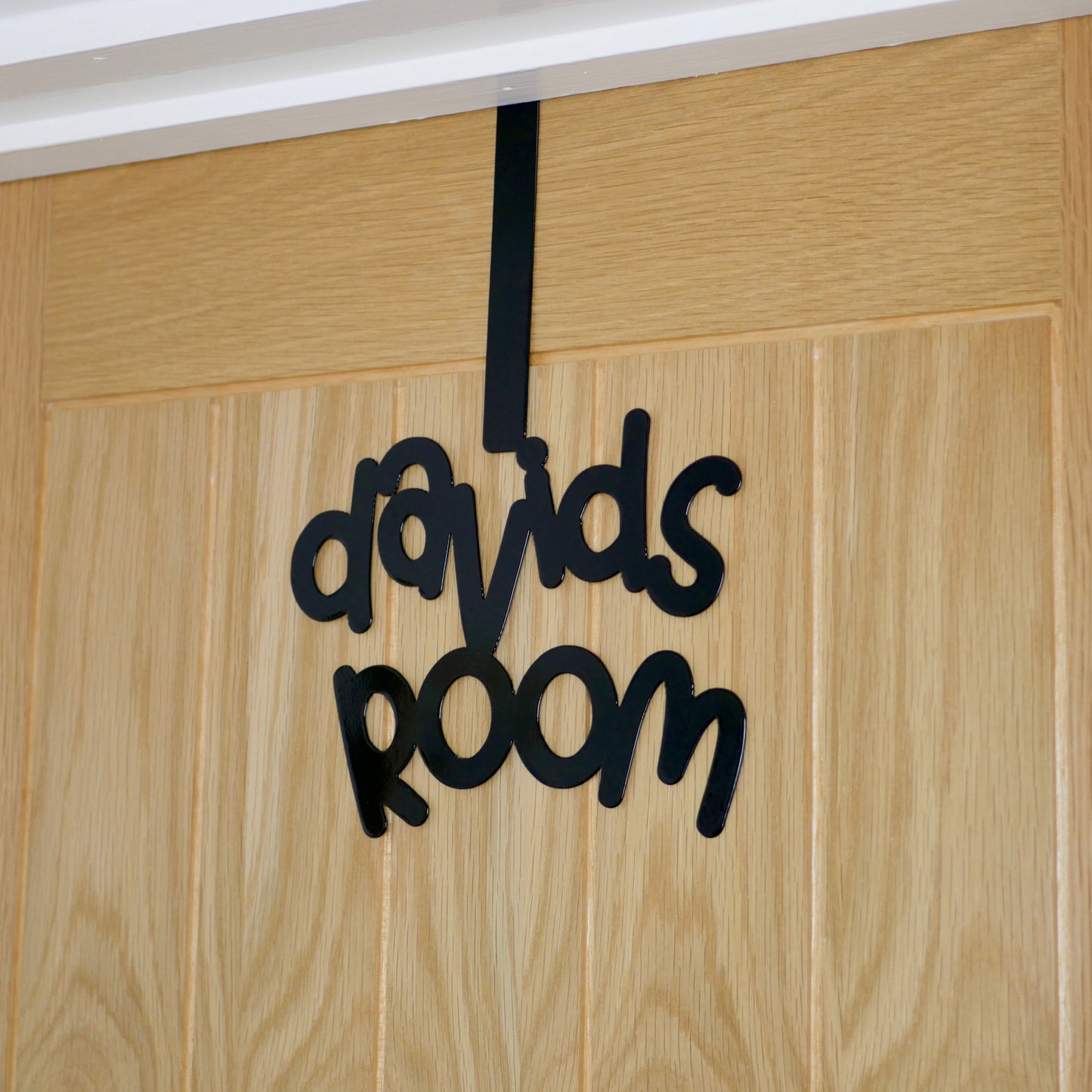 A wooden door with a black sign reading "davids room" that is in a cursive font. It is hung over the top of the door using an over door hook.