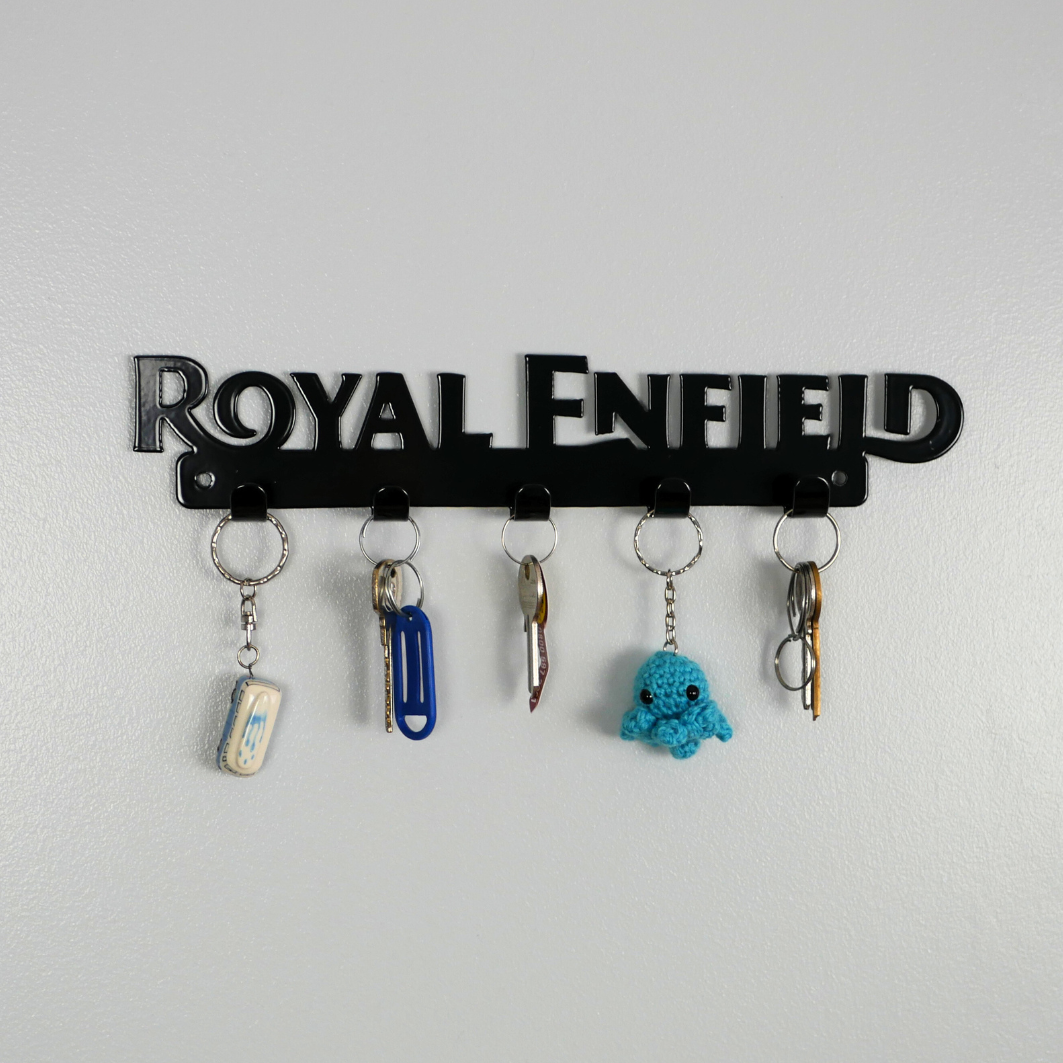 A close up of a black metal key rack in the shape of the Royal Enfield logo, it has 5 hooks and has two sets of keys and two keyrings hanging from them.
