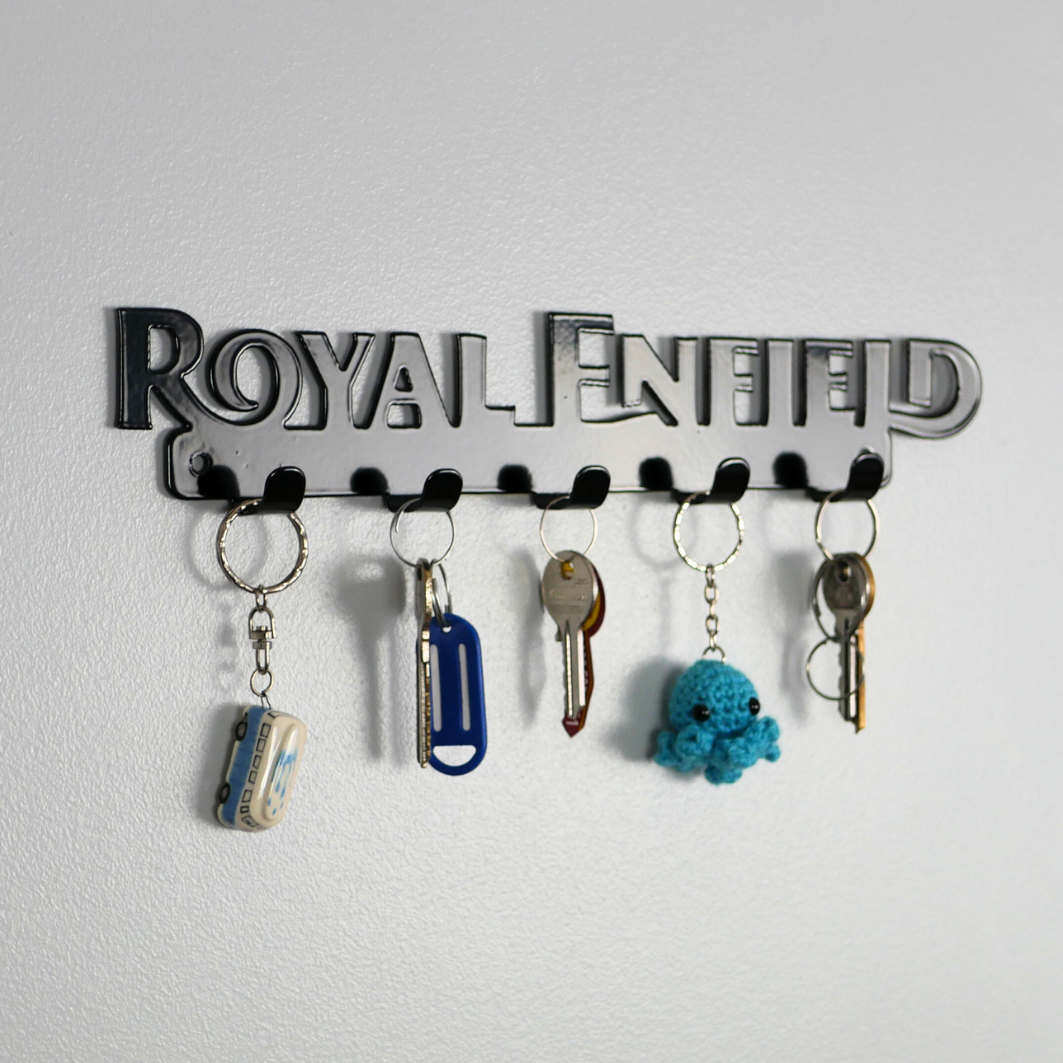 A left side view of a black metal key rack in the shape of the Royal Enfield logo, it has 5 hooks and has two sets of keys and two keyrings hanging from them.