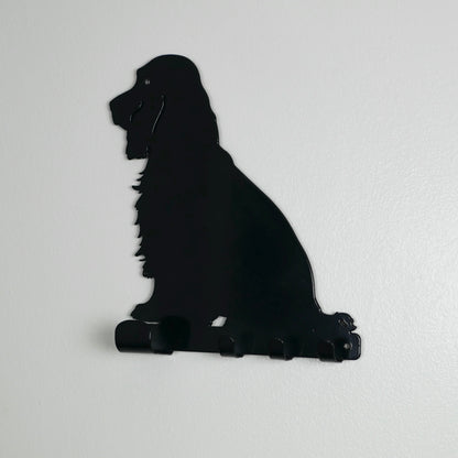 A side view of a black cocker spaniel dog key rack with one large hook and three smaller ones along the bottom. It is hung up on a white wall.