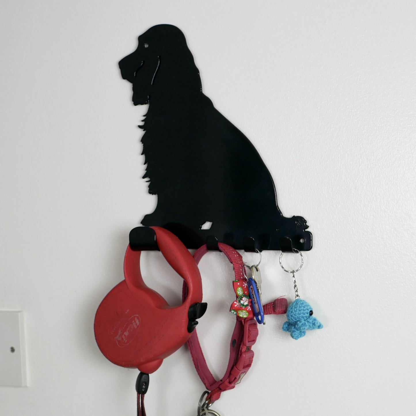 A black cocker spaniel dog key rack with one large hook and three smaller hooks along the bottom. Hanging from the hooks are a red dog lead, red dog collar, a set of  keys, and a keyring.