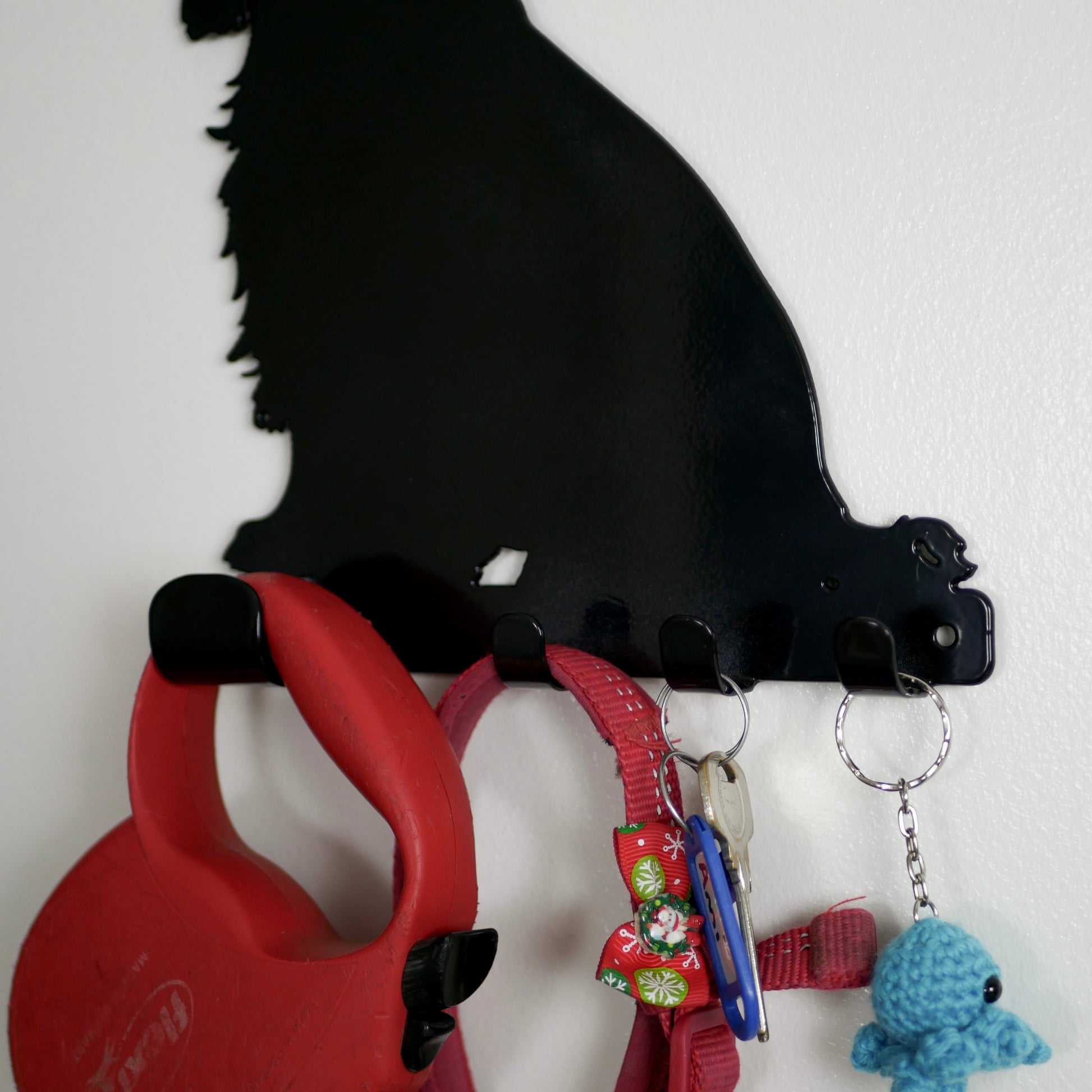 A close up image of a black cocker spaniel dog key rack with 1 large hook and 3 smaller hooks. Hanging from the hooks are a red dog lead, red dog collar, a set of keys, and a keyring.