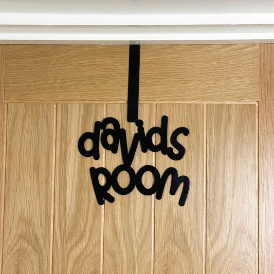 A wooden door with a black sign reading "davids room" that is in a cursive font. It is hung over the top of the door using an over door hook.