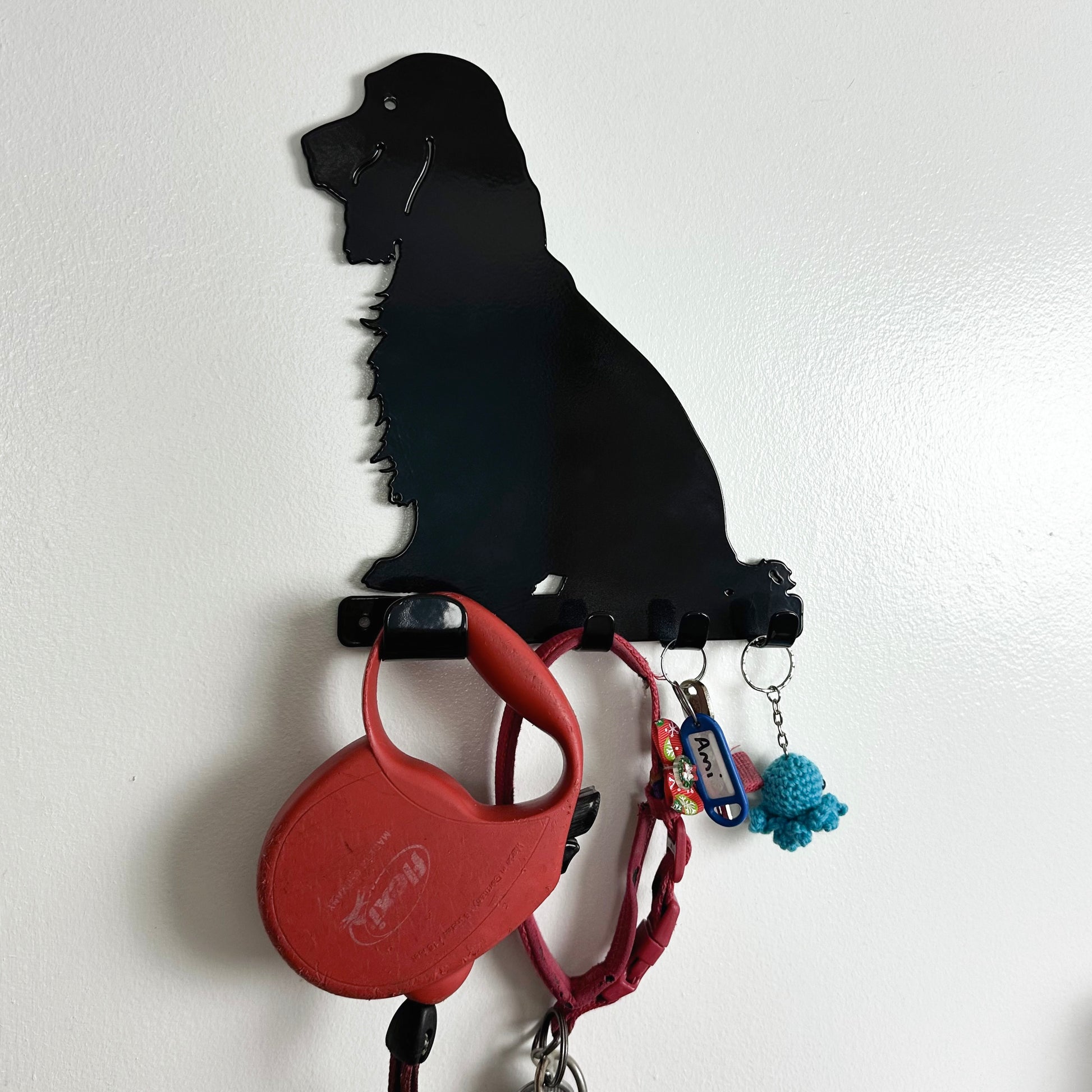 A black cocker spaniel dog key rack that is hanging on a wall, with a red dog lead, red dog collar, a set of keys, and a keyring hanging from the four hooks.