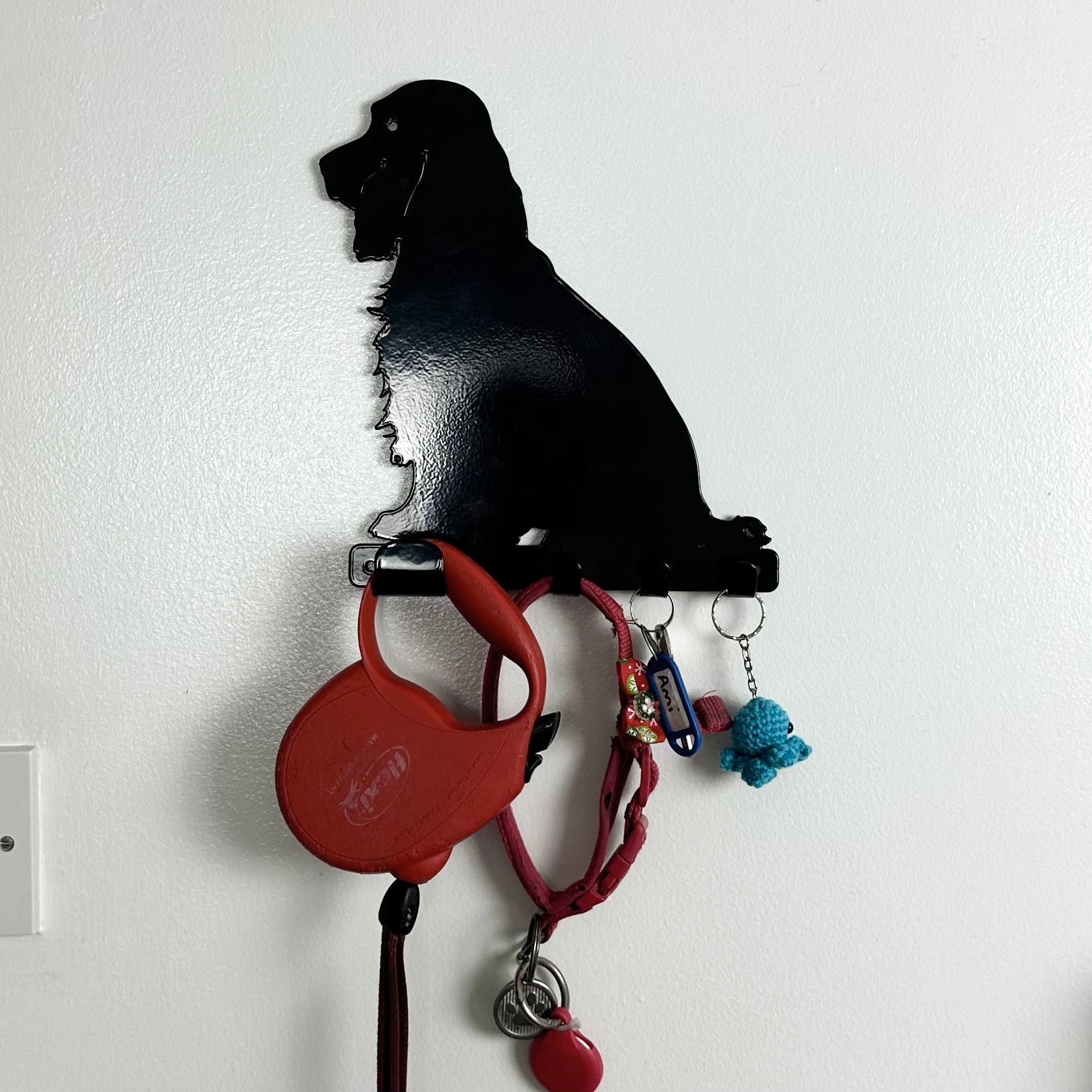 A wide shot of a black cocker spaniel dog key rack with one large hook and three smaller hooks along the bottom. Hanging from the hooks are a red dog lead, red dog collar, a set of  keys, and a keyring.