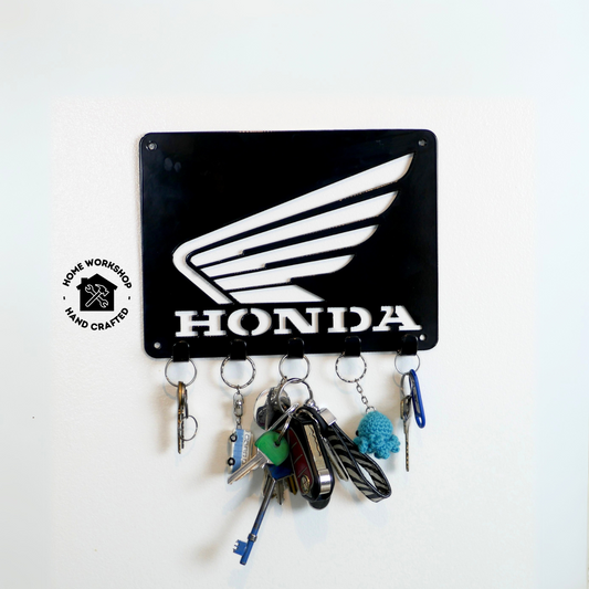 A black metal Honda logo hanging plaque with five small hooks along the bottom. Hanging from these hooks are two sets of keys, a set of car keys, and two keyrings.