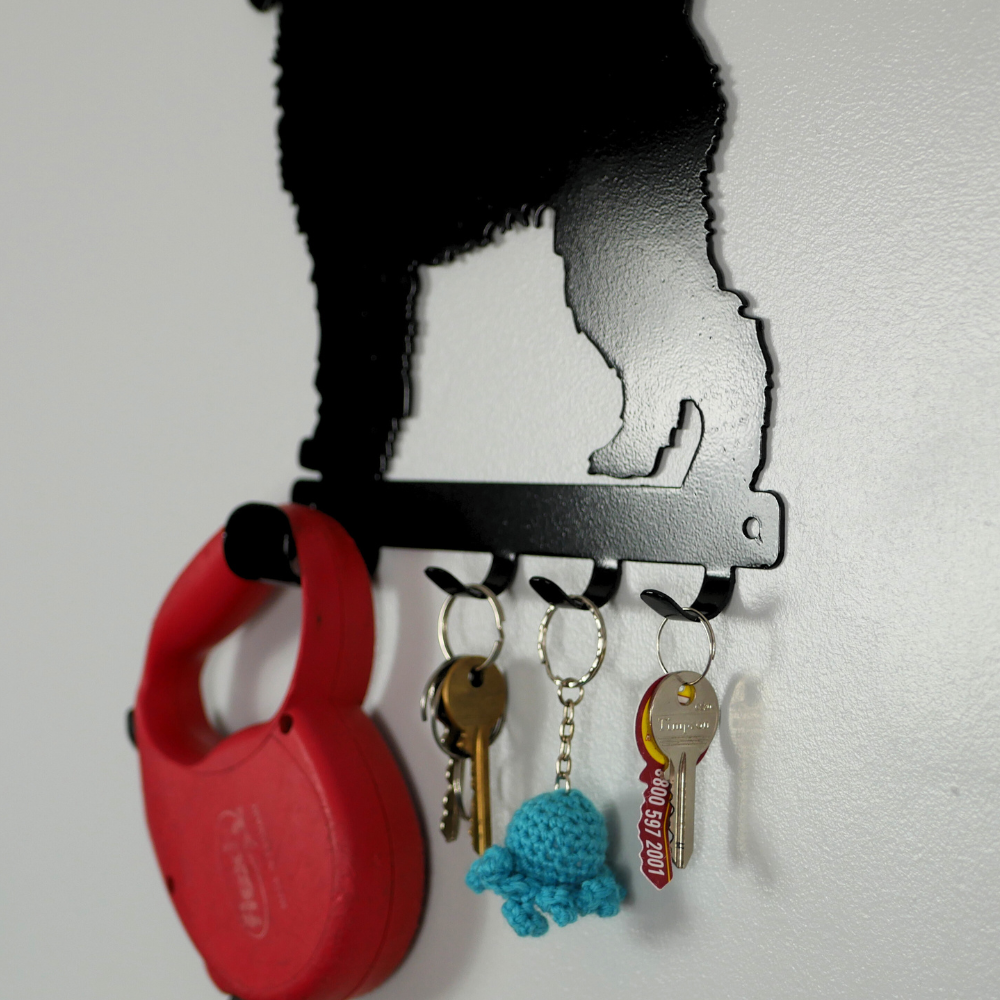 A close up photo of a cockerpoo dog shaped black metal dog lead and key rack, hanging from the four hooks are a dog lead, a keyring, and three keys.