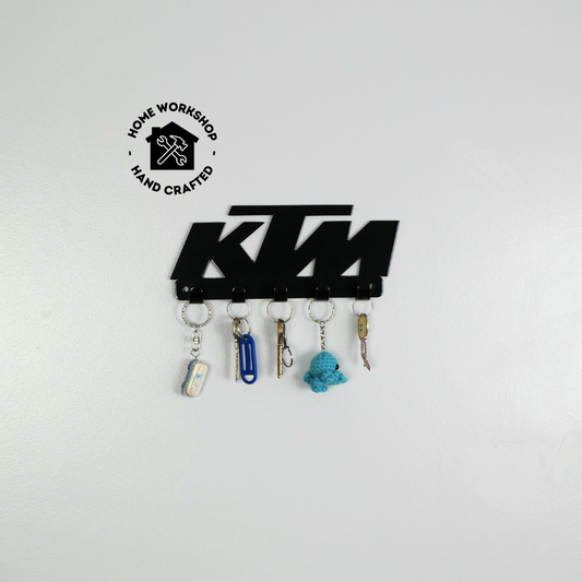 A black metal key rack in the shape of the KTM racing logo, it has 5 hooks and has two sets of keys and two keyrings hanging from them.