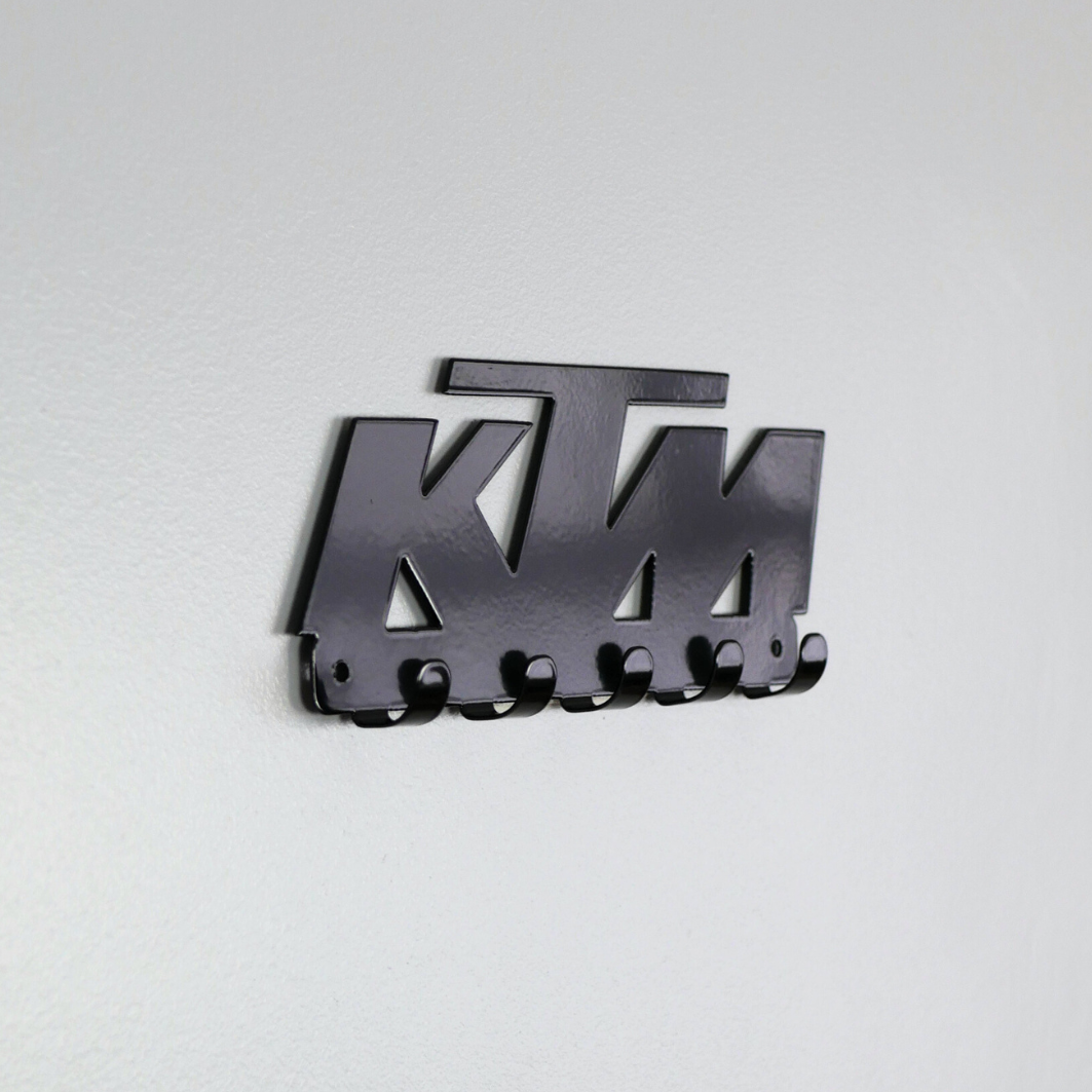A side view of a black metal key rack in the shape of the KTM racing logo, it has 5 hooks.