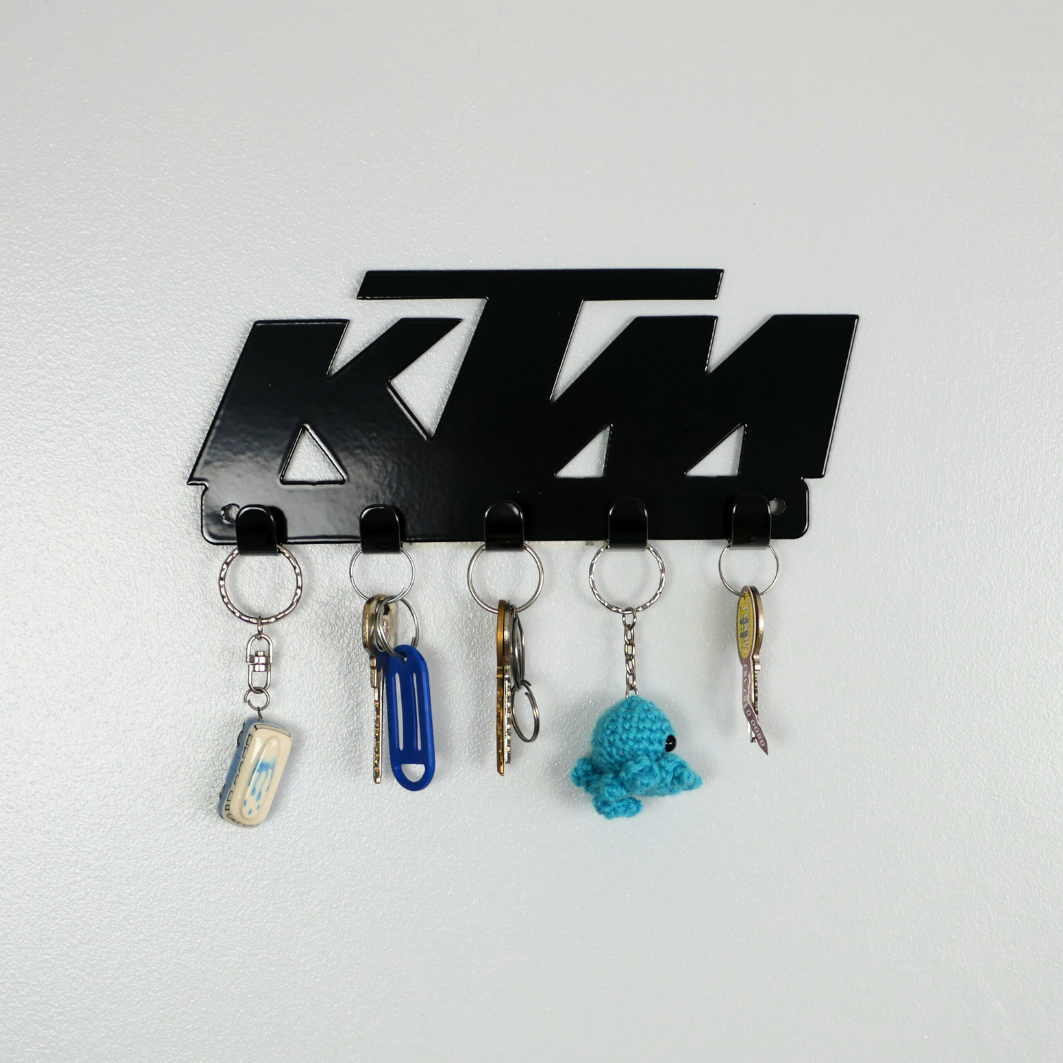 A close up of a black metal key rack in the shape of the KTM racing logo, it has 5 hooks and has two sets of keys and two keyrings hanging from them.