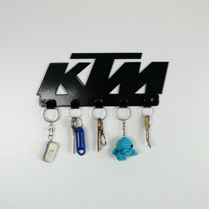 A close up of a black metal key rack in the shape of the KTM racing logo, it has 5 hooks and has two sets of keys and two keyrings hanging from them.