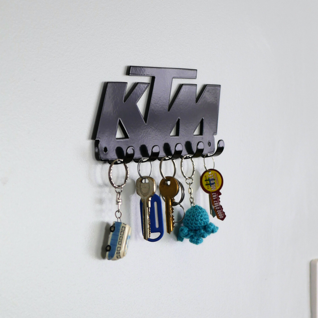 A side view of a black metal key rack in the shape of the KTM racing logo, it has 5 hooks and has two sets of keys and two keyrings hanging from them.