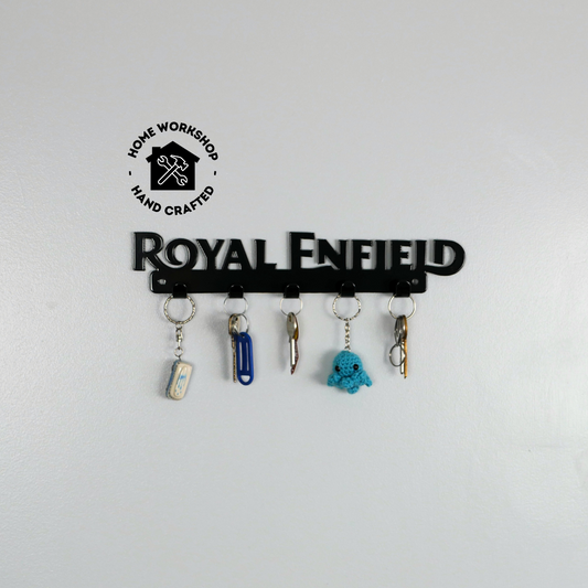 A black metal key rack in the shape of the Royal Enfield logo, it has 5 hooks and has two sets of keys and two keyrings hanging from them.