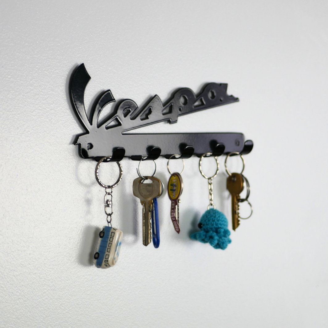 A left side angle of a black metal key rack in the shape of the Vespa logo, hanging from the 5 hooks are three sets of keys and two keyrings.