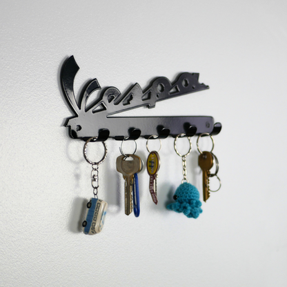 A left side angle of a black metal key rack in the shape of the Vespa logo, hanging from the 5 hooks are three sets of keys and two keyrings.