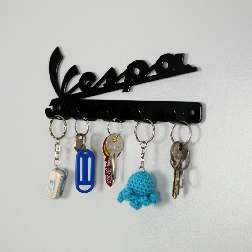 A right side angle of a black metal key rack in the shape of the Vespa logo, hanging from the 5 hooks are three sets of keys and two keyrings.