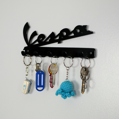 A right side angle of a black metal key rack in the shape of the Vespa logo, hanging from the 5 hooks are three sets of keys and two keyrings.