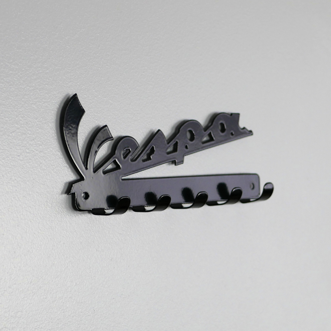 A left side angle of a black metal key rack in the shape of the Vespa logo, it has 5 hooks.