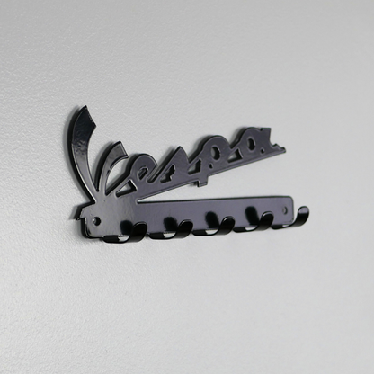 A left side angle of a black metal key rack in the shape of the Vespa logo, it has 5 hooks.